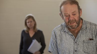 An older man with a distressed expression stands in the foreground, wearing a light blue patterned shirt. In the background, a woman holding papers looks on with a serious expression in a softly lit room.