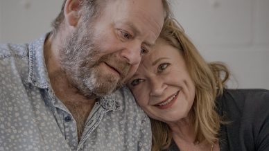 An older couple sits closely together, smiling warmly. The woman leans her head on the man's shoulder and they hold hands affectionately, conveying a sense of love and companionship.