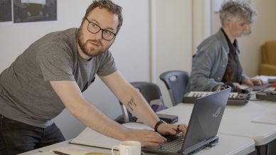 A man with glasses leans over a laptop on a table, looking up, with a notebook and coffee mug nearby. Another person works on a laptop and audio equipment in the background.