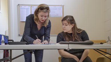 Two women work together at a desk, one standing and writing whilst the other sits and watches. A dog lies comfortably on a fluffy bed on the floor nearby. A noticeboard with papers hangs on the wall behind them.