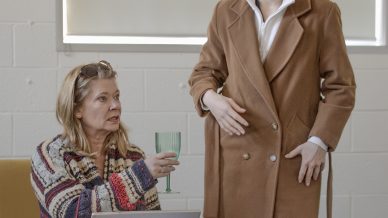 Two people indoors—one sitting on a yellow chair with a laptop, holding up a green glass, and the other standing, wearing a brown coat, looking at the seated person. A table with papers is in front of them.