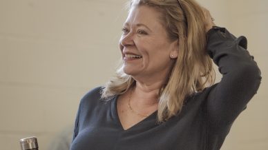 A woman with blonde hair smiles while holding a metal water bottle in one hand and touching her head with the other. She is wearing a dark long-sleeved top and glasses, standing indoors.