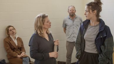 Four adults are in a bright room with white brick walls. Two women stand talking in the foreground; one holds a bottle. A man stands against the wall and another woman sits on a bench in the background.