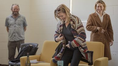 Three adults stand and laugh in a bright room. One woman puts on a colourful striped jumper near a yellow sofa, another woman in a brown coat smiles, and a man in glasses stands in the background. A bottle and glass sit on a table.