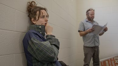 A woman in a checked jacket sits on the floor against a wall, looking thoughtful with her hand near her mouth. In the background, a man stands reading papers in a brightly lit, white-tiled room.