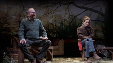 An older man and a younger woman sit on separate benches on stage, both looking pensive. The man holds papers and the woman sits with arms folded, a nature scene projected in the background.