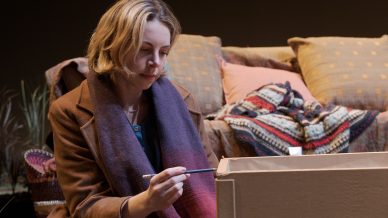 A woman with blonde hair in a brown coat sits in front of a cardboard box, writing on it with a pen. Behind her is a sofa with patterned cushions and a colourful blanket.