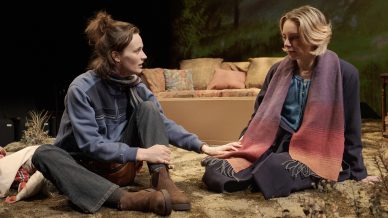 Two women sit on a stage floor covered in artificial grass, facing each other. One gently touches the other's hand. Behind them are a sofa and cushions, suggesting an indoor scene with a nature backdrop.
