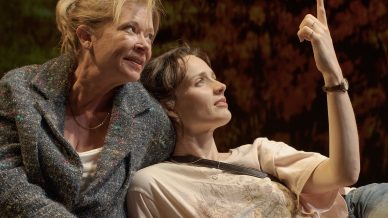 Two women sit closely together on the ground; one leans back against the other and points upward with a smile. They appear relaxed and content, surrounded by a warm, nature-inspired backdrop.