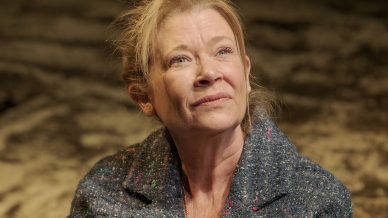 A middle-aged woman with blonde hair, wearing a blue speckled coat, sits on the ground with one arm across her chest, looking upwards with a thoughtful expression. The background is blurry and brown-toned.