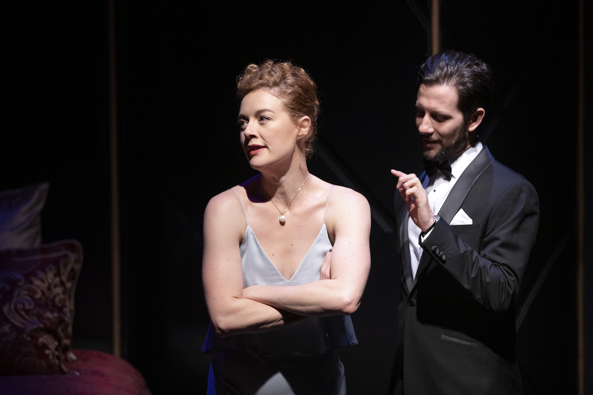 A woman in a satin dress stands with her arms folded, looking away, whilst a man in a black dinner jacket gestures with one hand behind her. The setting appears dramatic, with dim lighting and elegant decor.