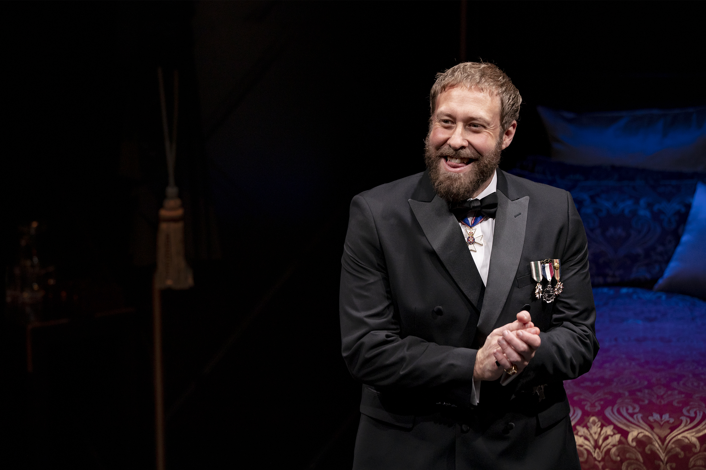A man with a beard, dressed in a formal black suit with medals on his chest, smiles and clasps his hands whilst standing on a dimly lit stage near a bed with ornate blue and purple bedclothes.