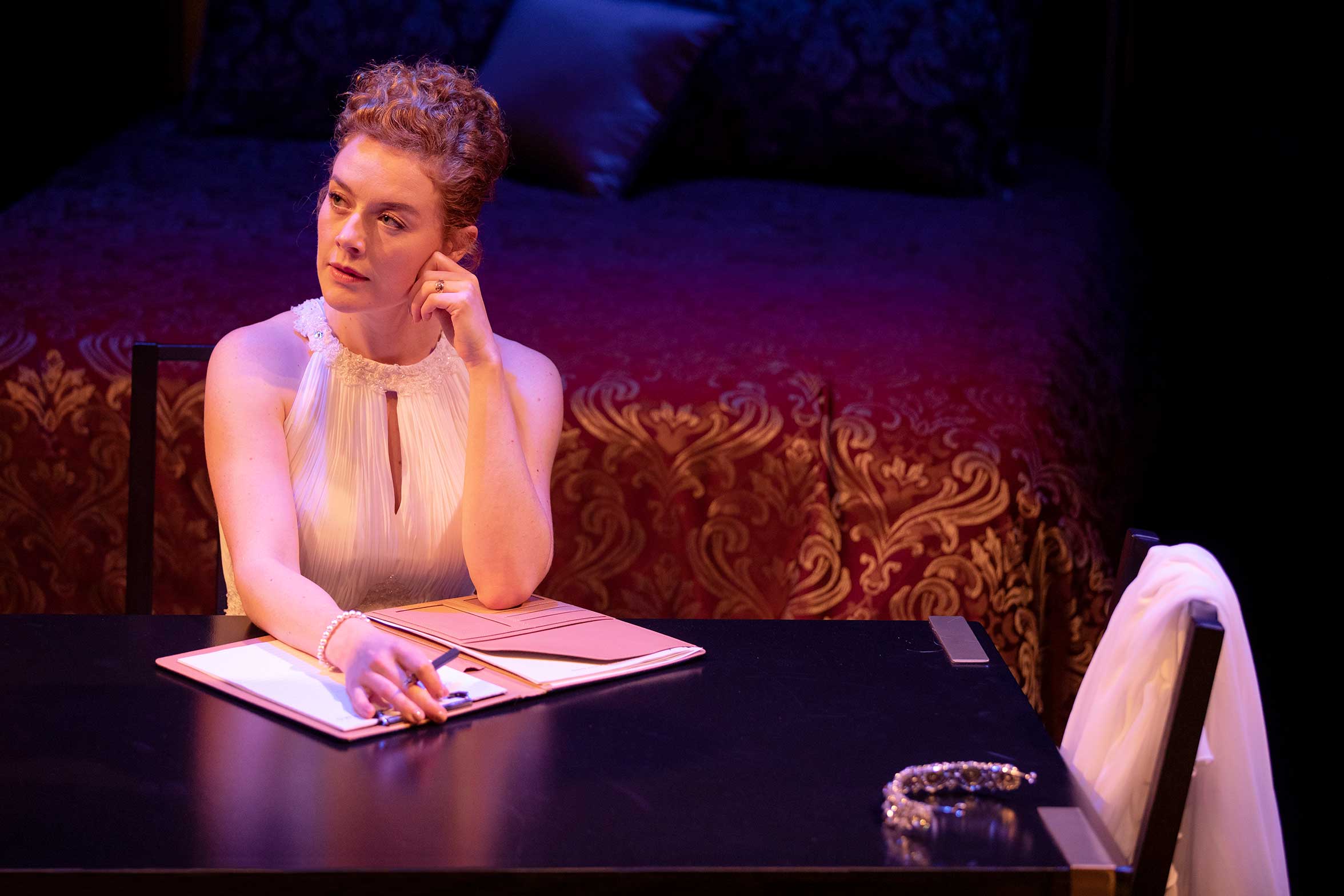 A woman in a white sleeveless dress sits at a table with a folder and pen, looking thoughtfully to the side. A bed with a red and gold patterned cover is in the background. A white jacket and jewellery rest on the table.