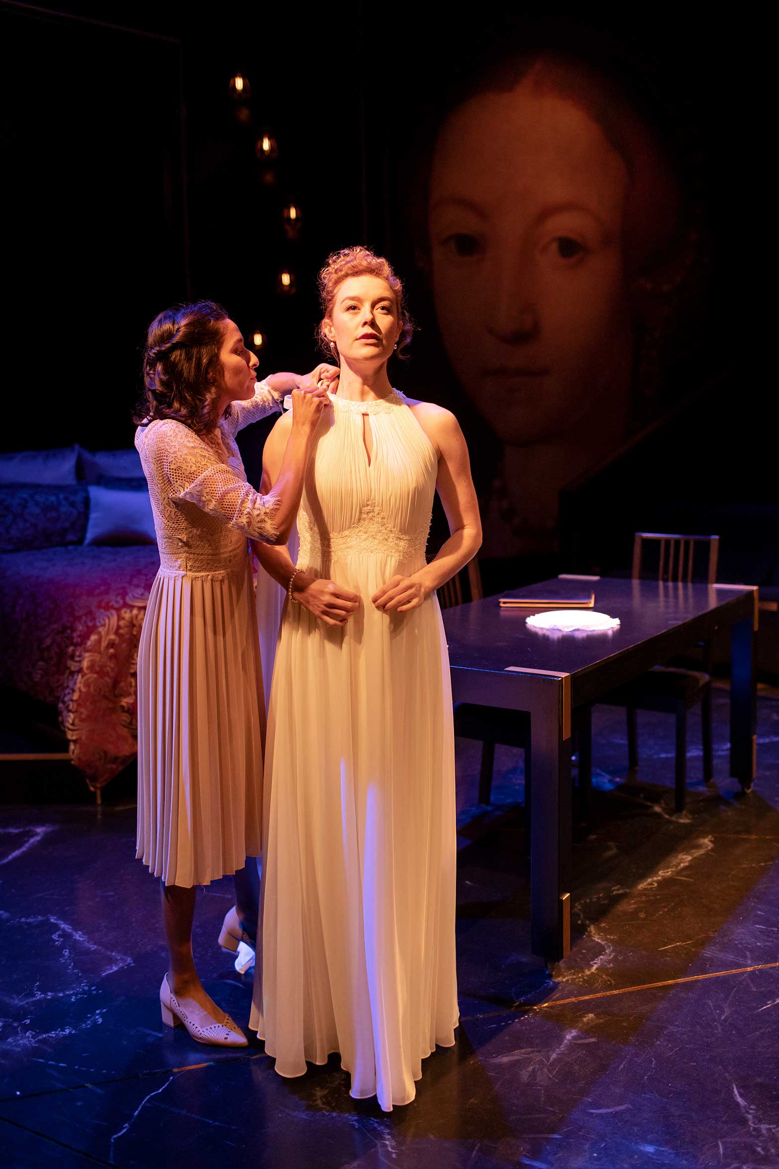 Two women stand on a dimly lit stage; one is helping the other adjust a long, elegant white dress. A large portrait of a woman's face is visible in the background, and the set includes a bed and a table with chairs.