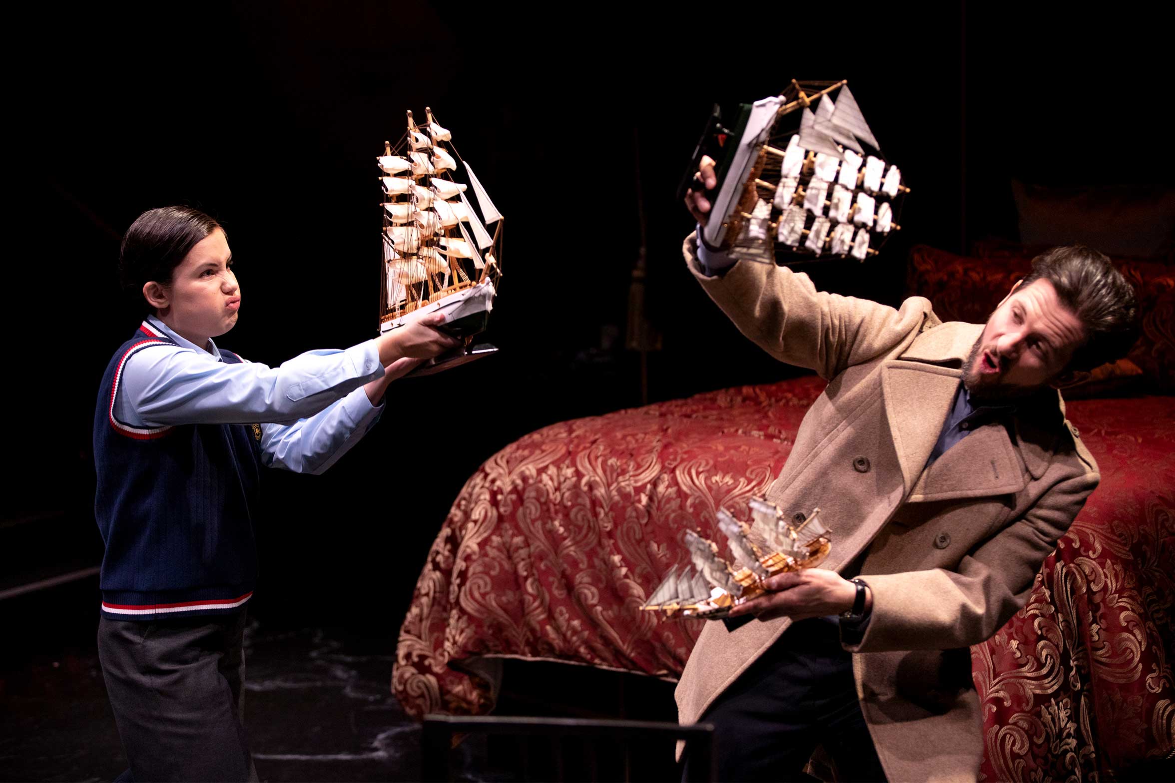 A child and an adult playfully pretend to battle with model ships, making animated faces. They stand in front of a large, ornate bed on a dark stage, suggesting a theatrical performance.