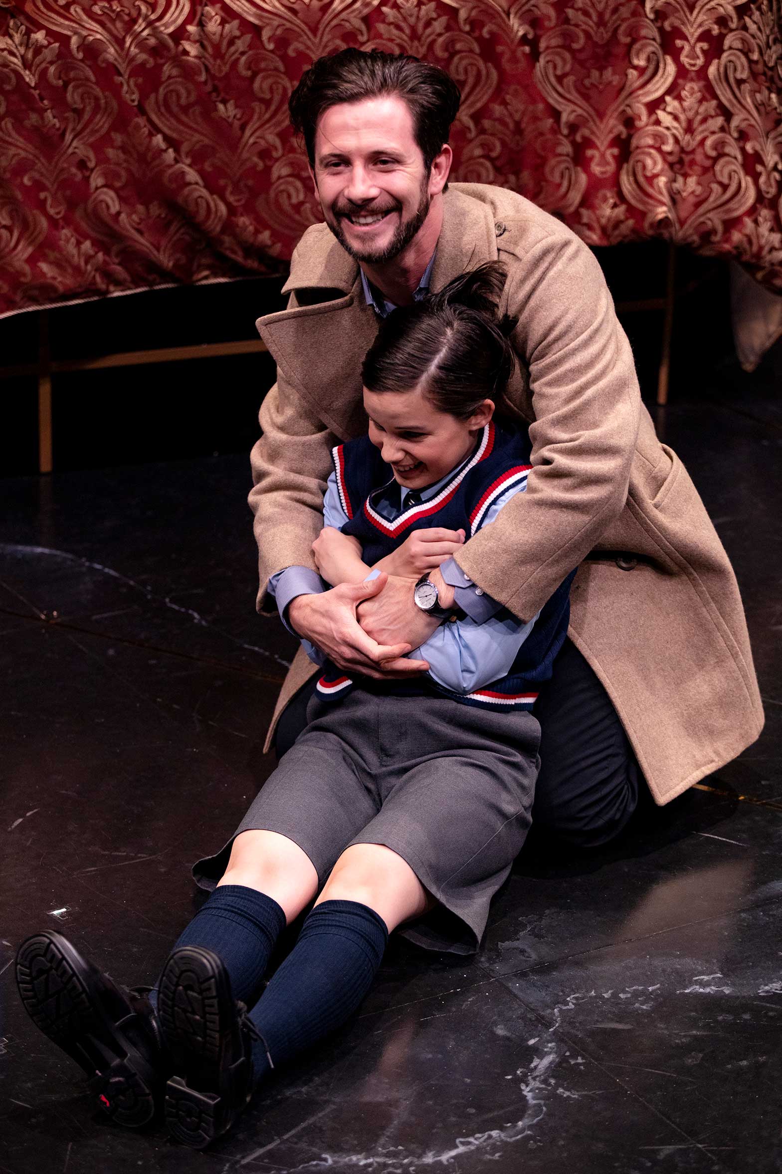 A smiling man in a tan coat sits on the floor, hugging a laughing child dressed in school uniform. They appear joyful, with a red patterned curtain in the background.