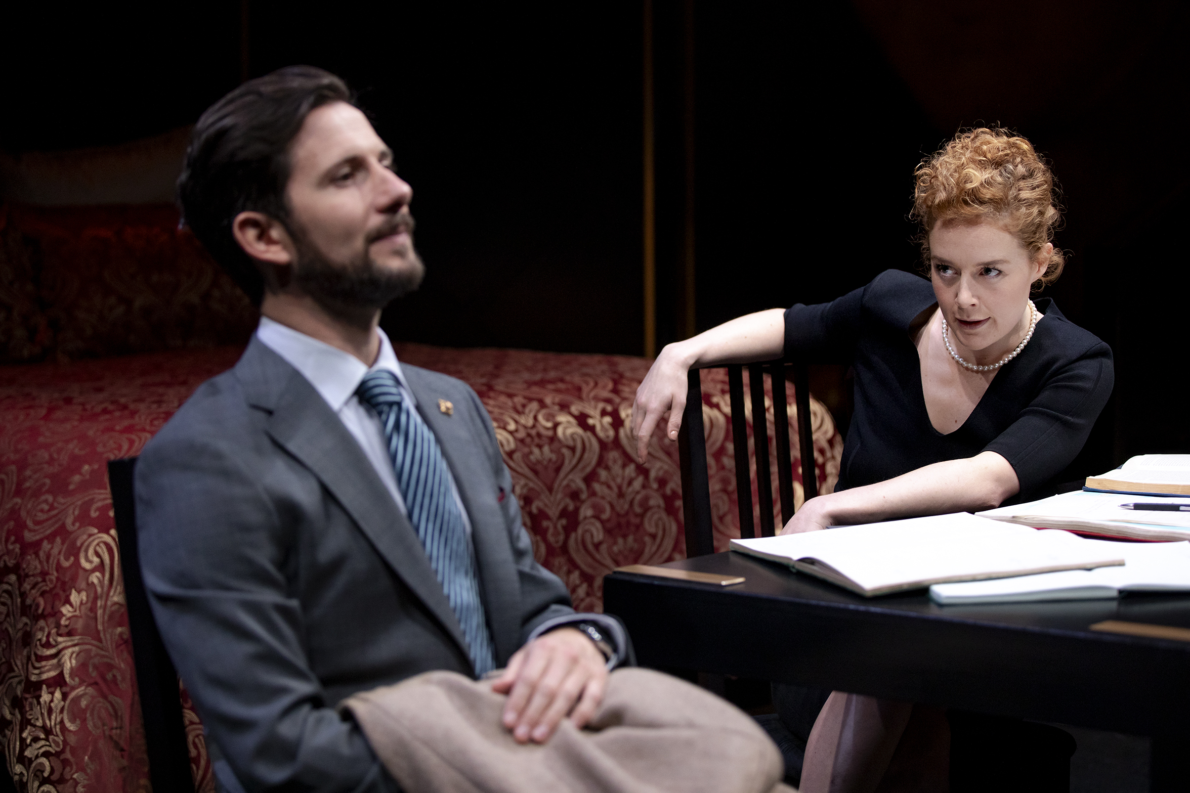 A man in a suit sits with a slight smile whilst a woman with curly red hair leans forward over a table covered in papers, looking at him intently. They appear to be in a dramatic conversation in a dimly lit room.