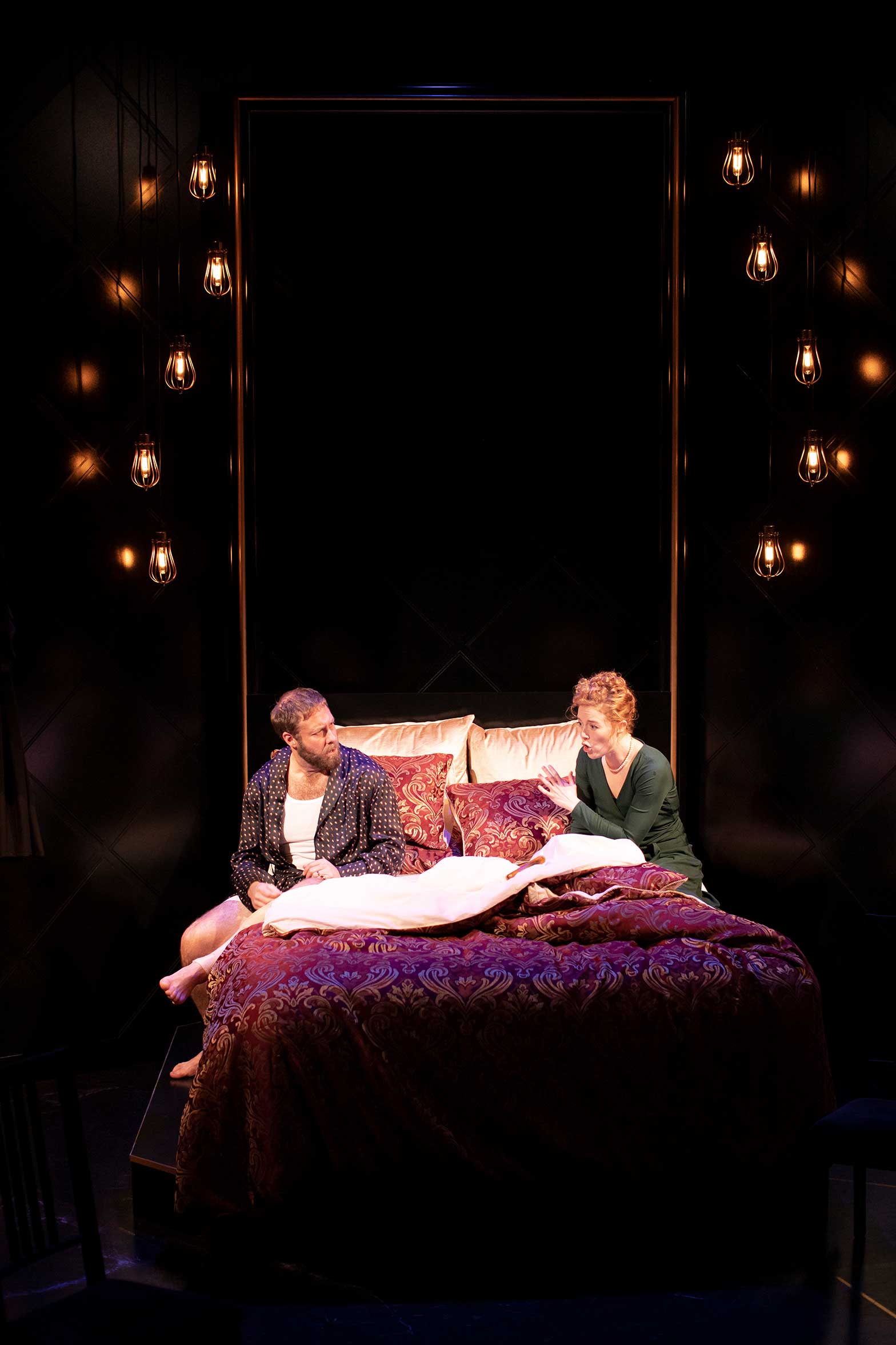 A man and a woman sit on an ornate, red and gold bed under warm hanging lights, facing each other in conversation. The setting appears theatrical, with dark surroundings highlighting the bed.
