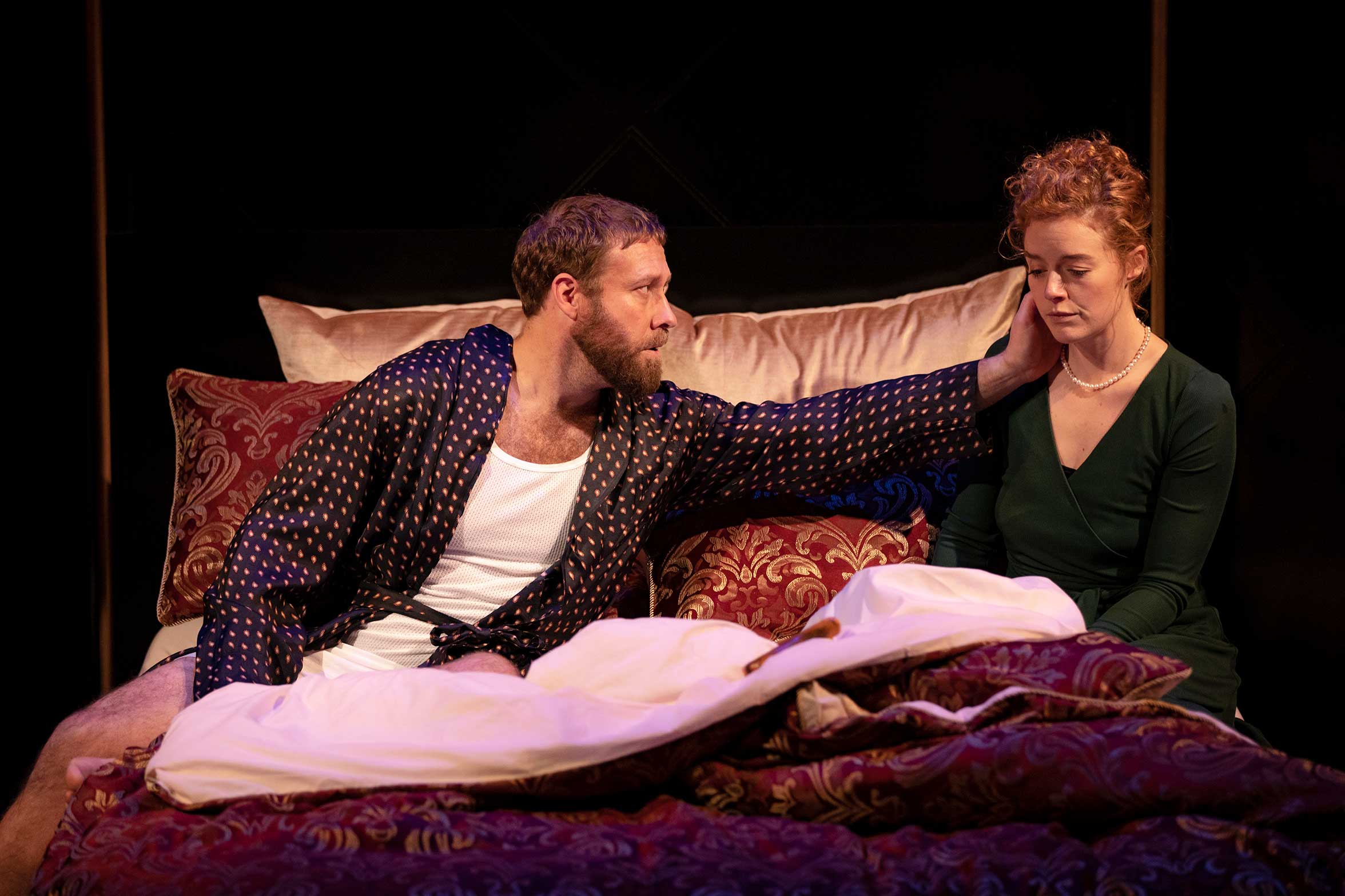 A man in a dressing gown sits on a bed, gently touching the face of a woman in a green dress beside him. The woman looks down, appearing thoughtful or sad. They are surrounded by patterned cushions and bedding.