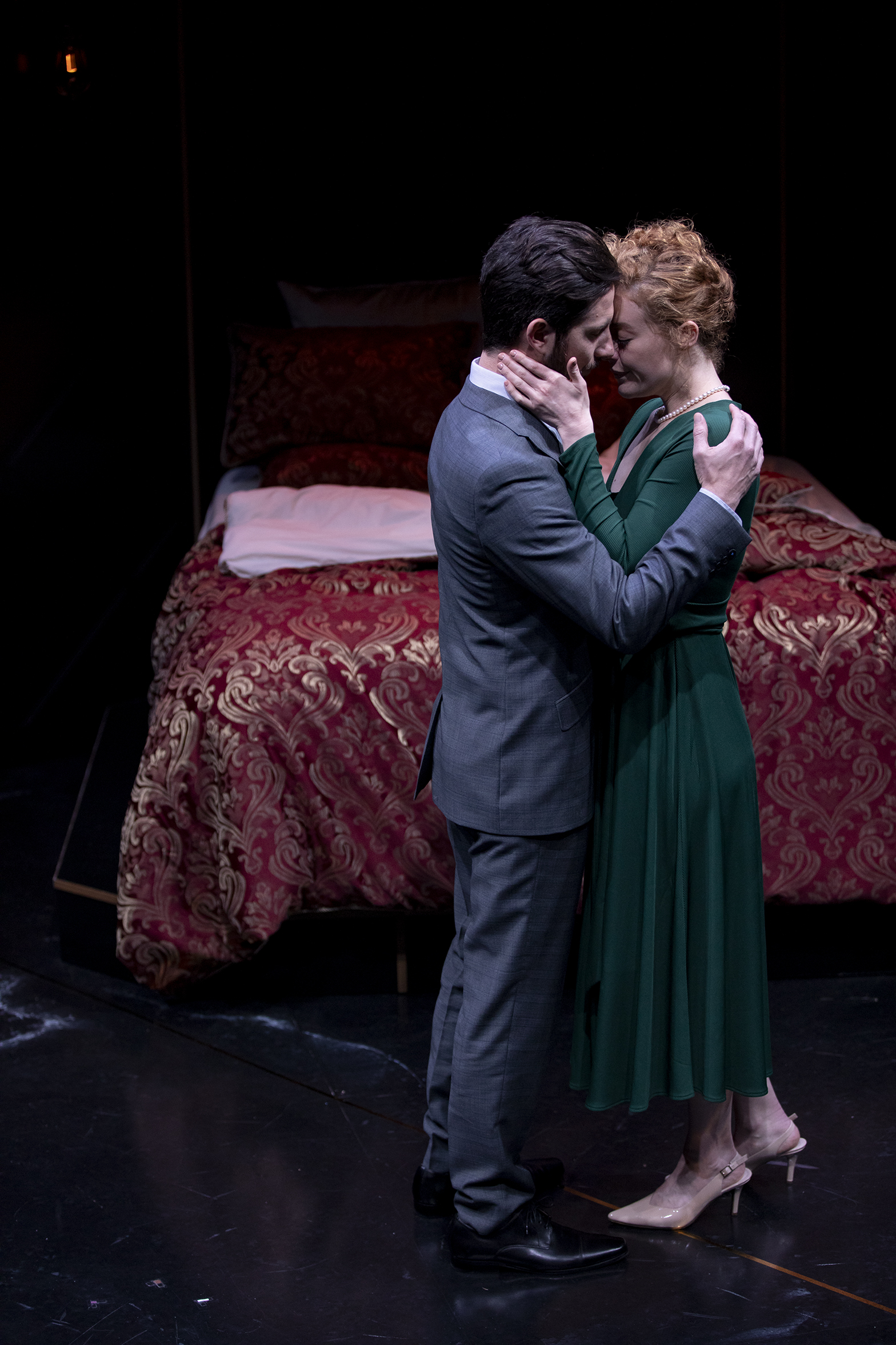 A man in a grey suit and a woman in a green dress stand close together, touching foreheads intimately, in front of a bed with a red patterned bedspread on a dimly lit stage.