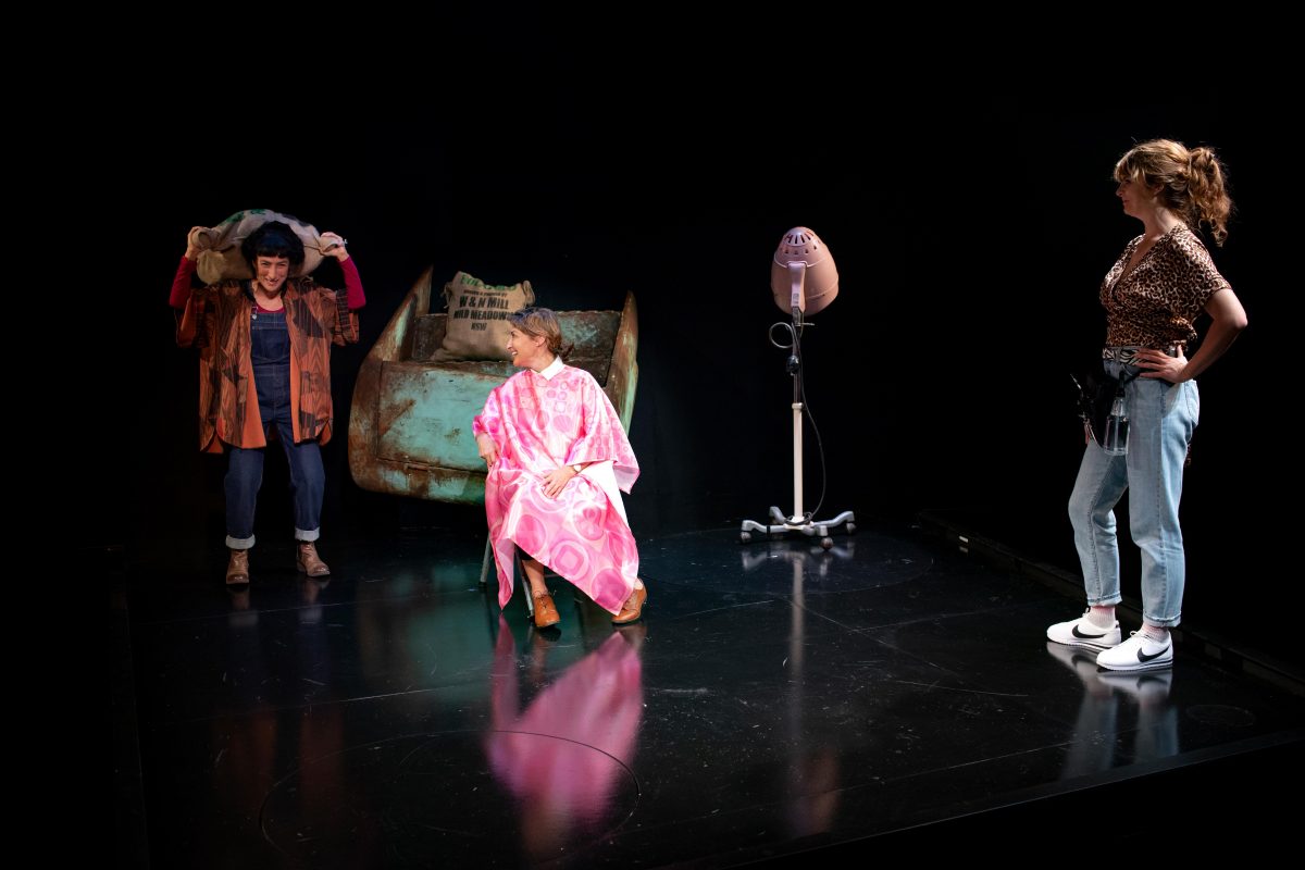 Three women on a stage: one stands smiling and holding a bag over her head, another sits in a salon chair wearing a pink gown, and the third stands watching with hands on hips near a vintage hairdryer.