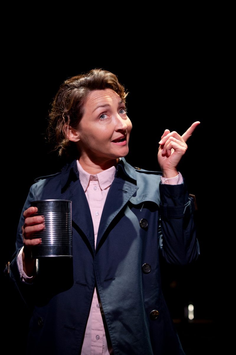 A woman in a dark trench coat holds a tin in one hand and points upwards with the other, looking engaged and expressive against a dark background.