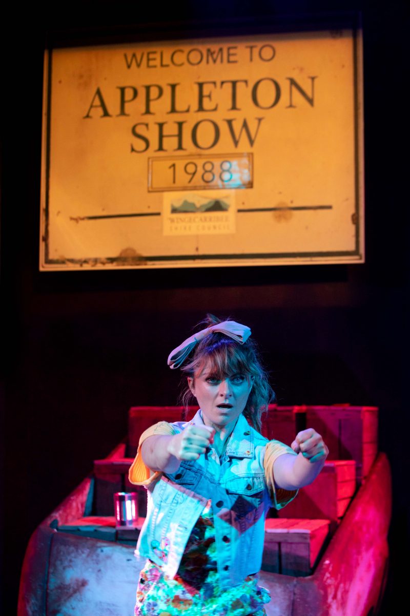 A young woman in colourful clothing and a bow headband gestures as if driving. Behind her is a sign reading Welcome to Appleton Show 1988 and a stack of wooden crates, all under dramatic stage lighting.