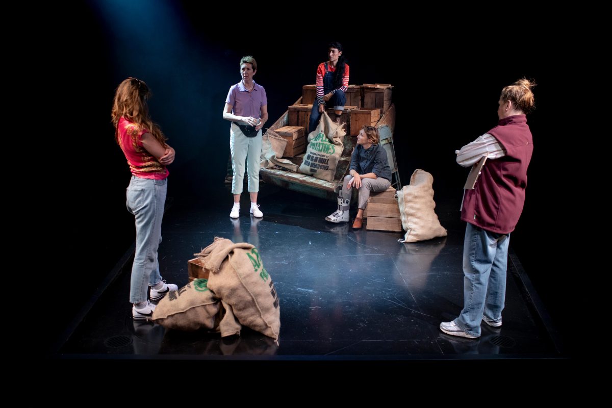 Five people stand in a circle around hessian sacks and crates on a dimly lit stage, engaged in conversation. The set suggests a warehouse or storage area, with wooden boxes stacked in the background.