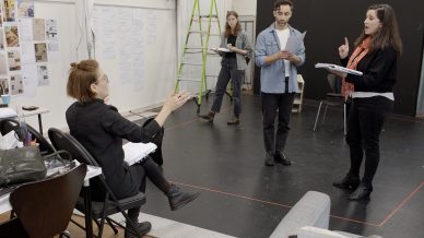 Four people in a rehearsal room: one seated woman gestures while talking to three standing actors with scripts, one of whom is speaking and gesturing. A ladder, notes on the wall, and a table with bags are visible in the background.
