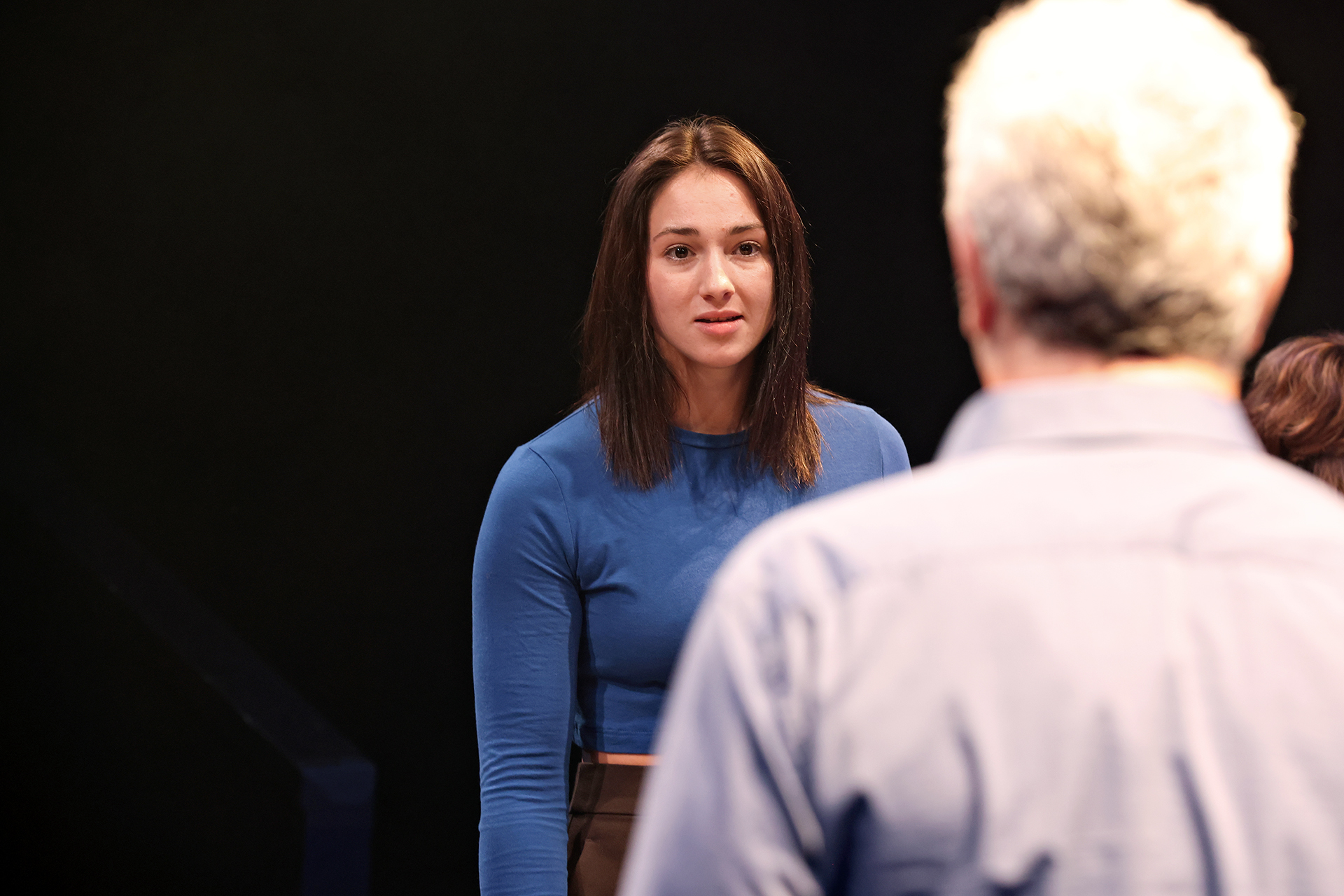A young woman with straight brown hair, wearing a blue long-sleeved shirt, faces an older man with grey hair, seen from behind, in a dimly lit setting. She appears to be listening or responding to him.