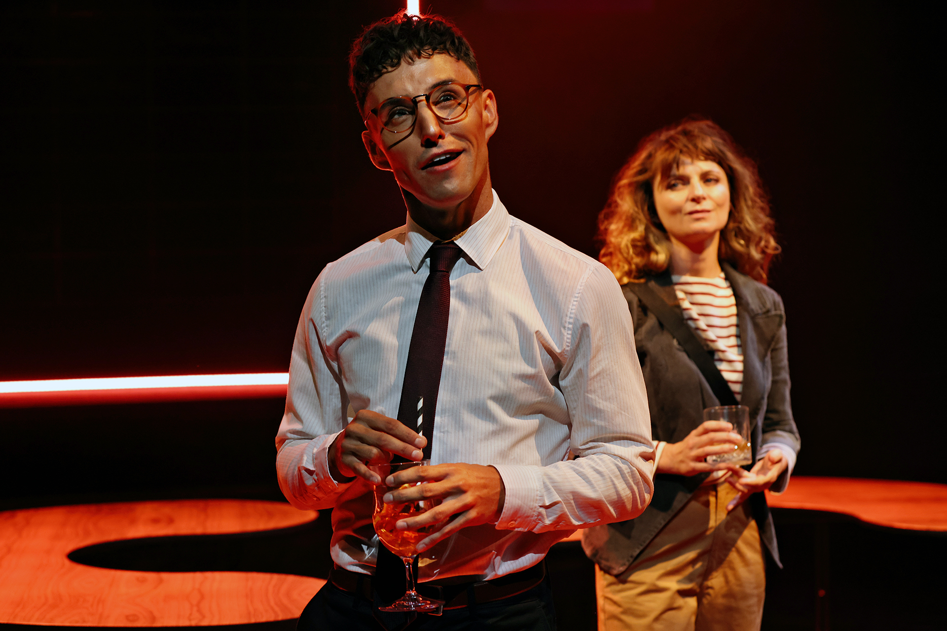A man in glasses and a tie holds a drink and looks upwards, while a woman in a blazer and striped shirt stands behind him, also holding a drink. They are on a dimly lit stage with red lighting in the background.