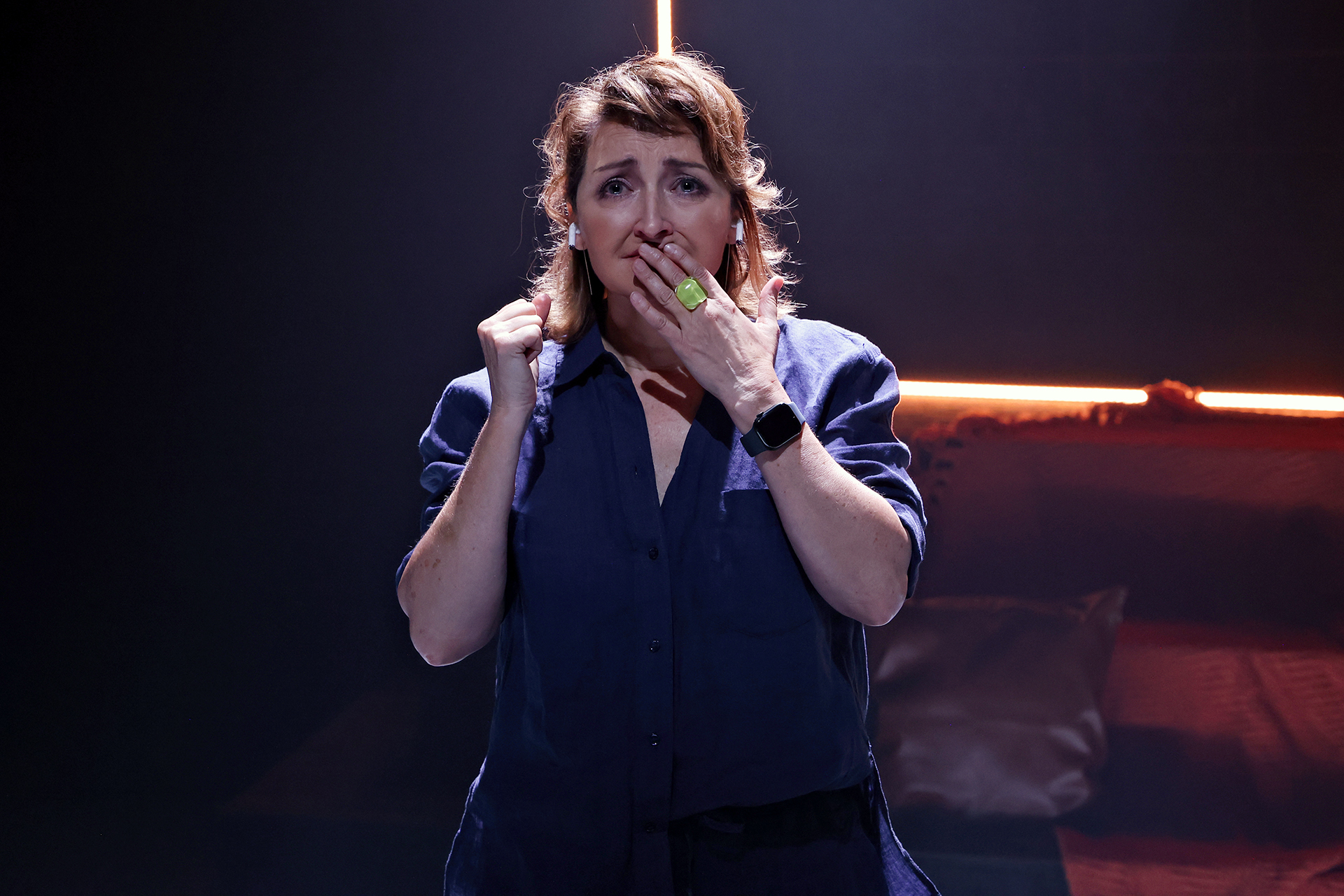 A woman stands in dramatic lighting with a worried expression, one hand raised to her mouth and the other clenched. She wears a dark shirt and an Apple Watch, with an illuminated background behind her.