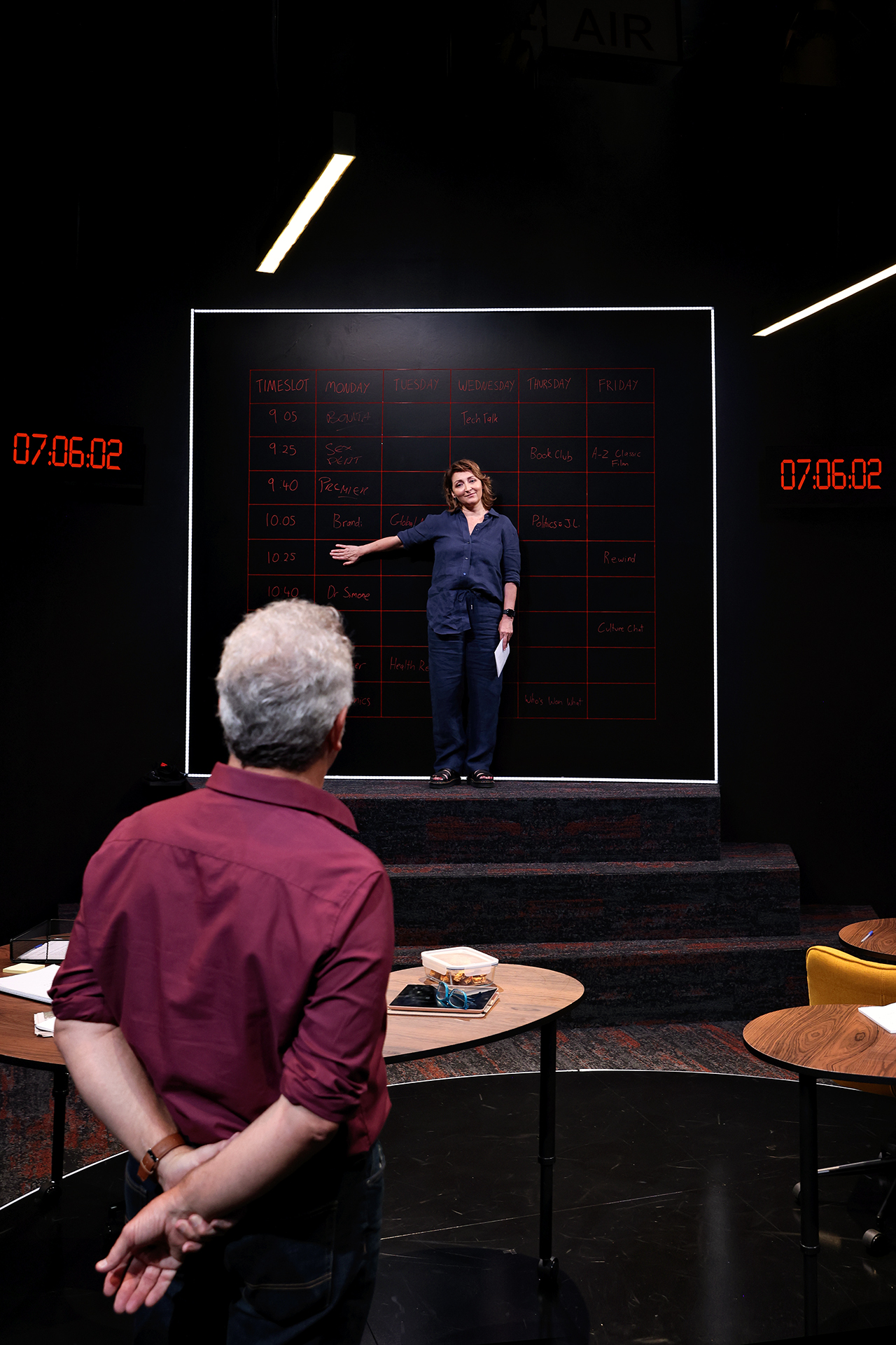 A person stands in front of a large blackboard with a weekly timetable written on it, pointing to the board, while another person stands in the foreground facing them. Two digital clocks display 07:06:02 on either side.