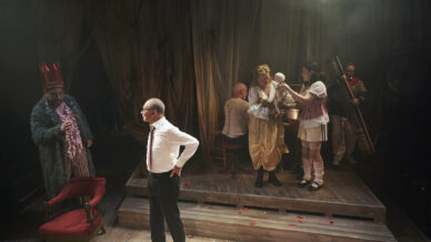 A theatrical stage scene with five actors in costume: one in a crown and fur coat, one in a white shirt standing apart, and three interacting near a chair. The stage is dimly lit with rose petals on the floor.