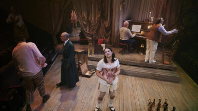 A woman in period costume stands centre stage, surrounded by men in historical attire. One man plays a piano, while others interact and move props on a wooden stage set with draped curtains and scattered objects.