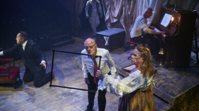 A theatrical scene on stage shows five actors; one kneels with arms out, another stands looking through a rectangular frame held by a woman in a white blouse, whilst a man plays piano and another stands in the background.