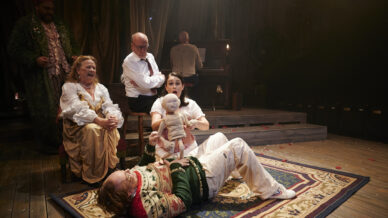 Four actors in theatrical costumes perform on stage. Two sit on chairs, one kneels holding a doll, and another lies on a rug. A pianist plays in the background. The scene is dramatic and warmly lit.