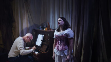 A man plays the piano while a woman in a pink corset and white dress stands in front of him, singing passionately. Dim, moody lighting and draped curtains set a dramatic stage atmosphere.