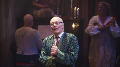 An elderly man in a patterned dressing gown stands centre stage, clasping his hands and looking upward. Behind him, one person plays a piano and another stands holding a recorder. The scene is warmly lit with candlelight.