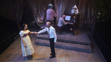 A man in a white shirt holds the hand of a woman in a white dress on a wooden stage, while three other people are in the background near a piano, including one dressed as a clown. The scene is set in a dimly lit theatre.