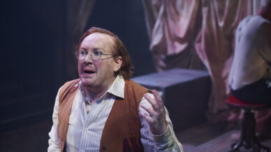 A man with glasses and reddish hair, wearing a striped shirt and brown waistcoat, gestures expressively on stage with dramatic lighting and draped curtains in the background.