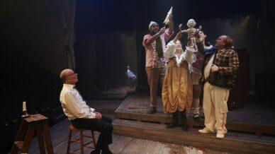 Five actors perform on a dimly lit stage. Four stand together, holding up a puppet baby, while one sits on a stool watching them. The stage floor is wooden with scattered red petals.