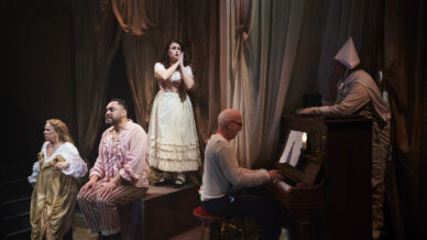 Five actors perform on stage in period costumes; one plays the piano, two sit and sing, one stands singing passionately, and one in a white hat leans on the piano. The setting is draped with soft, flowing fabric.