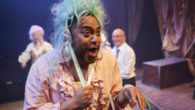 A person in a light pink ruffled costume and teal wig performs expressively on stage; two people in period costumes, one with white hair, are blurred in the background under dramatic lighting.