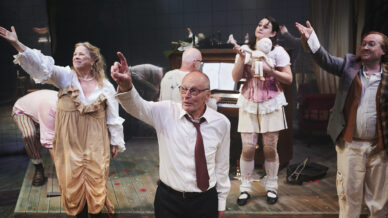 Five actors on stage in dramatic poses; one elderly man in a white shirt stands front and centre. Behind him, others wear eclectic costumes, including one woman holding a doll, with a cluttered set and scattered rose petals.