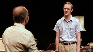 Two men are indoors, having a serious conversation. One man with red hair, glasses, and a tucked-in shirt faces forward, while the other, seen from behind, gestures with his hands. A lamp and sofa are visible in the background.