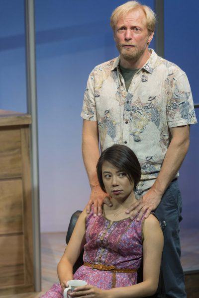 A man with blond hair stands behind a seated woman with short dark hair, resting his hands on her shoulders. The woman, wearing a pink patterned dress, holds a white mug and looks to the side. They are indoors with a blue background.