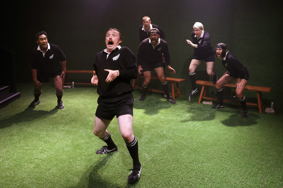 Six people in black rugby kits perform on a green artificial turf stage. The person at the front has an exaggerated, surprised expression, while the others in the background appear animated and engaged in the scene.