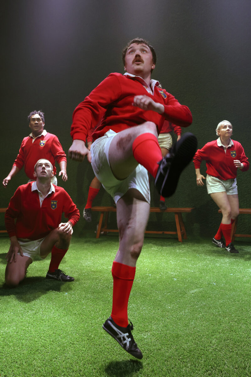 A group of rugby players in red shirts and white shorts play on green turf. One player is mid-kick, raising his leg high, while others watch intently in the background.