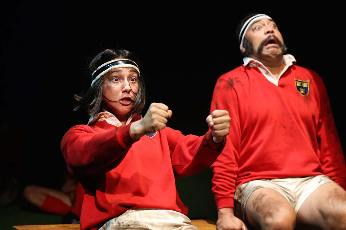 Two people in red rugby kits and headbands sit side by side. The person in front is gripping imaginary reins, looking intense, whilst the person behind has a dramatic, wide-eyed expression. Both have mud stains on their clothes.